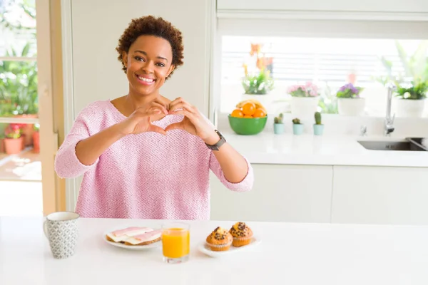 Young african american woman eating breaksfast in the morning at home smiling in love showing heart symbol and shape with hands. Romantic concept.