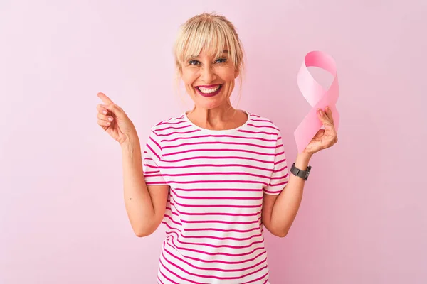 Middle age woman holding cancer ribbon standing over isolated pink background very happy pointing with hand and finger to the side