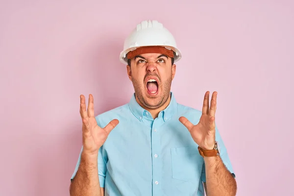 年轻的建筑师头戴安全帽 站在孤立的粉色背景之上 疯狂而疯狂地大喊大叫 带着咄咄逼人的表情和手臂 沮丧的概念 — 图库照片