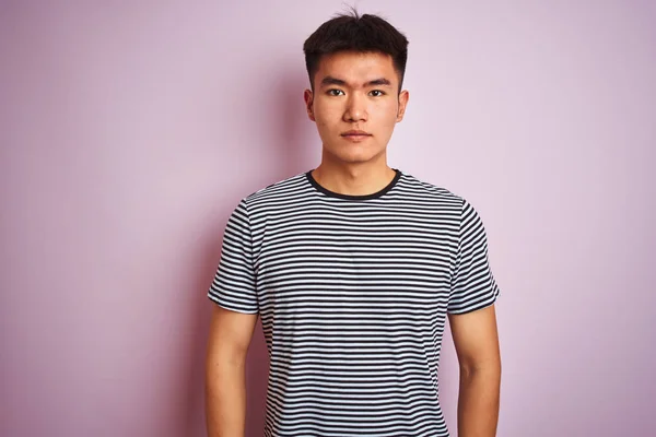 青白いピンクの背景の上に立つストライプのTシャツを着たアジアの若い中国人男性は 真剣な表情を浮かべていた カメラを見てシンプルで自然な — ストック写真