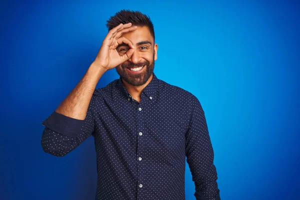 孤立した青い背景の上に立つシャツを着た若いインドのエレガントな男は 手の笑顔でOkジェスチャーを行い 幸せな顔で指を通して目を見ます — ストック写真