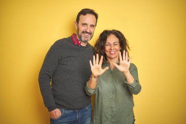 İzole sarı arka plan üzerinde güzel orta yaş çift gösteren ve parmak numarası on ile işaret kadar güven ve mutlu gülümseyen.