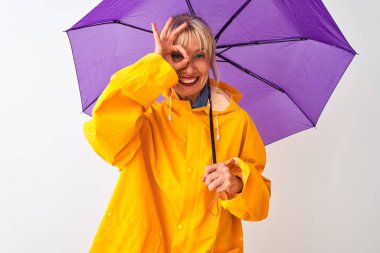 Orta yaş kadın izole beyaz arka plan üzerinde yağmur ceket ve mor şemsiye giyen mutlu yüzü parmakları ile bakarak göz üzerinde el ile ok işareti yapıyor gülümseyerek