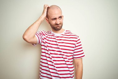 Young bald man with beard wearing casual striped red t-shirt over white isolated background confuse and wondering about question. Uncertain with doubt, thinking with hand on head. Pensive concept.