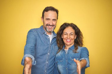 Güzel orta yaş çift birlikte izole sarı arka plan üzerinde tebrik ve karşılama olarak dostça sunan el sıkışma gülümseyerek üzerinde duran. Başarılı iş.