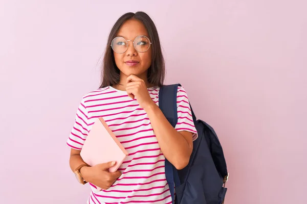 Chinese student woman wearing glasses backpack holding book over isolated pink background serious face thinking about question, very confused idea