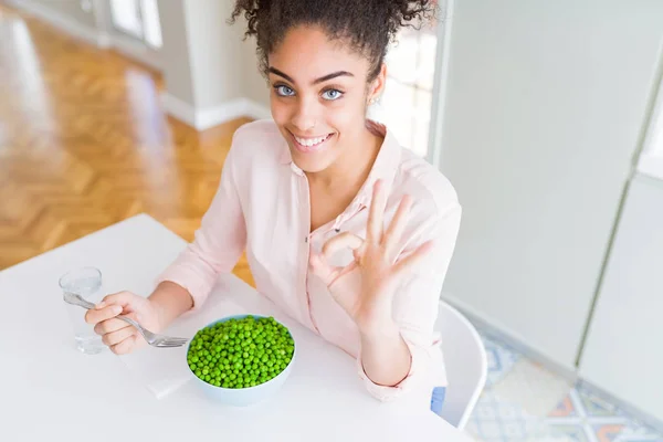 Young african american girl eating healthy green peas doing ok sign with fingers, excellent symbol