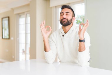 Yakışıklı, beyaz süveter giyen, evde rahat ve gülümseyen, gözleri kapalı meditasyon yapan, parmaklarıyla hareket eden bir İspanyol. Yoga konsepti.