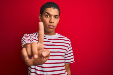 İzole edilmiş kırmızı arka plan üzerinde çizgili tişört giyen genç yakışıklı Arap adam parmağını kaldırmış ve kızgın bir ifade sergiliyor.