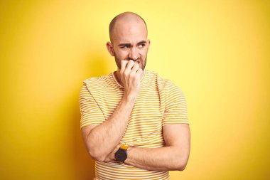 Sakallı genç kel adam sarı izole arka plan üzerinde rahat çizgili t-shirt giyen stresli ve ağız tırnak ısırma elleri ile sinir görünümlü. Anksiyete sorunu.