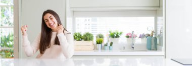 Geniş açı gülümseyen ve başarı için çığlık evde çok mutlu ve heyecanlı kaldırdı, silah ile kazanan jest yapıyor beyaz masada oturan güzel genç kadın resim. Kutlama kavramı.