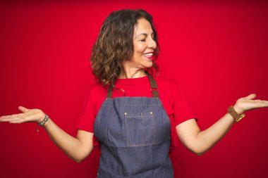 Orta yaş yaşlı kadın kırmızı izole arka plan üzerinde önlük üniforma giyen gülümseyerek her iki elaçık avuç içi gösteren, sunan ve reklam karşılaştırma ve denge