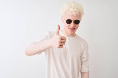 Genç albino sarışın adam izole beyaz arka plan üzerinde t-shirt ve güneş gözlüğü giyen el ile jest kadar mutlu başparmak yapıyor. Başarı göstererek kameraya bakan ifadeyi onaylama.
