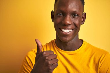 African american man wearing casual t-shirt standing over isolated yellow background happy with big smile doing ok sign, thumb up with fingers, excellent sign