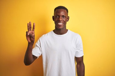 Beyaz tişört giyen genç afro-amerikan adam izole sarı arka plan üzerinde duran gösteren ve parmakları ile işaret ederken kendinden emin ve mutlu gülümseyerek üç numara.
