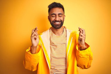 İzole sarı arka plan gesturing parmak üzerinde duran yağmurluk giyen genç Hintli adam umut ve gözleri kapalı gülümseyerek geçti. Şans ve batıl kavram.