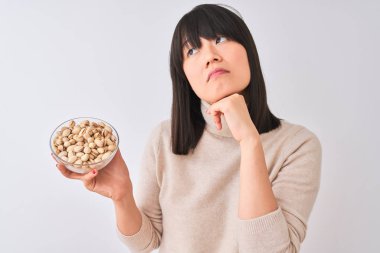 Genç güzel Çinli kadın izole beyaz arka plan üzerinde antep fıstığı ile kase tutan ciddi yüz soru hakkında düşünme, çok karışık bir fikir