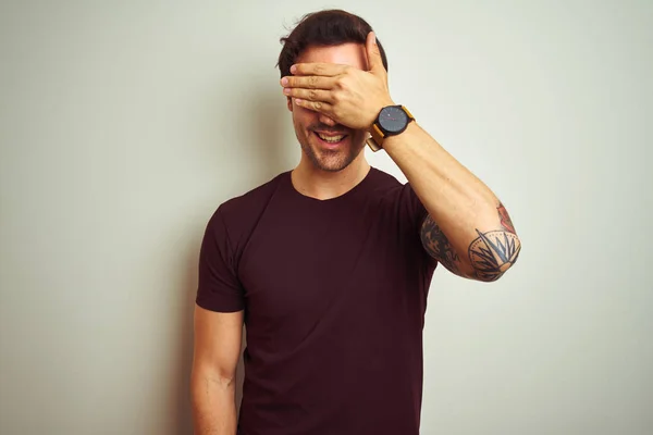 Young handsome man with tattoo wearing purple casual t-shirt over isolated white background smiling and laughing with hand on face covering eyes for surprise. Blind concept.