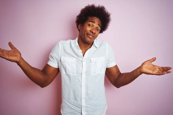 Young american man with afro hair wearing white shirt standing over isolated pink background clueless and confused expression with arms and hands raised. Doubt concept.