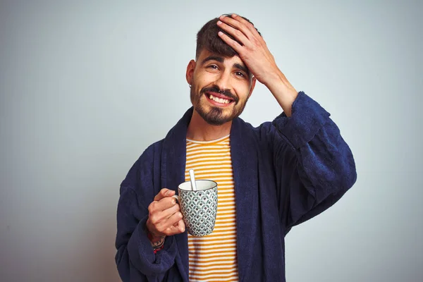 Young man wearing bathrobe drinking cup of coffee standing over isolated white background stressed with hand on head, shocked with shame and surprise face, angry and frustrated. Fear and upset for mistake.