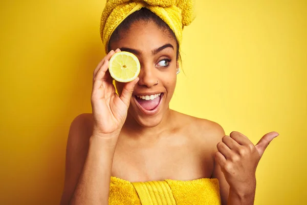 シャワーの後にタオルを着たアフロの女性は 孤立した黄色の背景の上にスライスレモンを保持し 幸せな顔で側に親指で示しています — ストック写真