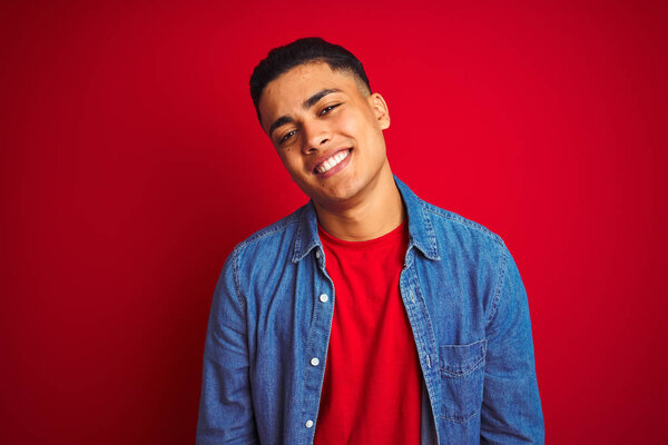 Young brazilian man wearing denim shirt standing over isolated red background looking away to side with smile on face, natural expression. Laughing confident.