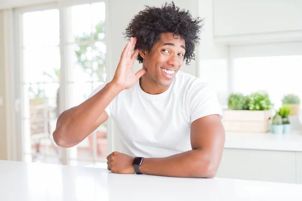 Young african american man wearing casual white t-shirt sitting at home smiling with hand over ear listening an hearing to rumor or gossip. Deafness concept.