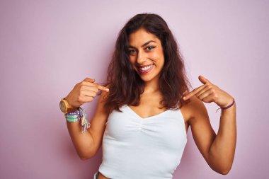 İzole pembe arka plan üzerinde duran beyaz t-shirt giyen genç güzel kadın yüzünde gülümseme ile emin görünümlü, parmakları gururlu ve mutlu kendini işaret.