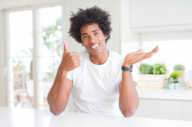 Genç afro-amerikan adam evde oturan rahat beyaz t-shirt giyen avuç içi el gösteren ve başparmak ile ok jest yapıyor, mutlu ve neşeli gülümseyerek