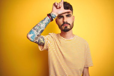 Çizgili t-shirt giyen dövme giyen genç adam izole sarı arka plan üzerinde duran ezik jest alay ve hakaret yapıyor alnına parmakları ile insanların alay yapma.