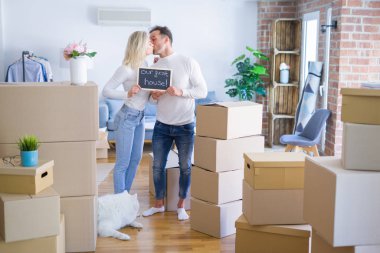 Karton kutular etrafında yeni evde öpüşme mesajı ile tahta tutan ayakta köpek ayakta genç güzel çift