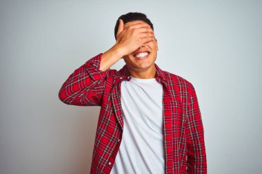 Kırmızı gömlek giyen genç brezilyalı adam izole beyaz arka plan üzerinde duran gülümseyerek ve sürpriz için gözleri kaplayan yüzünde el ile gülüyor. Kör kavramı.