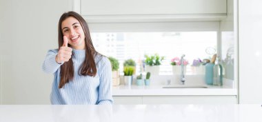 Evde beyaz masada oturan güzel genç kadının geniş açılı resmi el ile jest kadar mutlu başparmak yapıyor. Başarı gösteren kameraya bakarak ifadeyi onaylama.