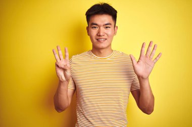Genç asyalı Çinli adam izole sarı arka plan üzerinde duran t-shirt giyen gösteren ve parmakları ile işaret ederken emin ve mutlu gülümseyerek sekiz numara.
