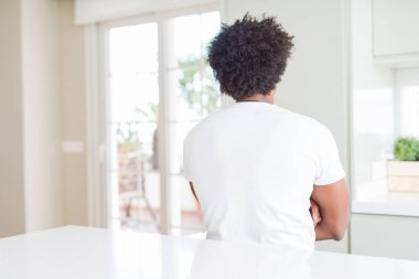 Evde oturan rahat beyaz tişört giyen genç afro-amerikan adam çapraz kollarıyla geriye doğru bakıyor