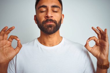 İzole beyaz arka plan üzerinde duran t-shirt giyen genç hintli adam rahatlayın ve gözleri parmakları ile meditasyon jest yapıyor kapalı gülümseyerek. Yoga konsepti.