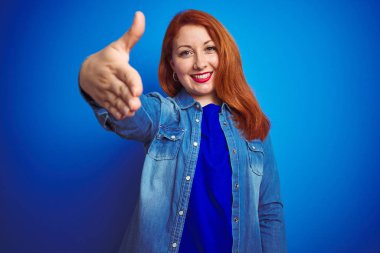 Mavi izole edilmiş arka planda duran kot tişörtlü, güzel, kızıl saçlı bir kadın selamlaşma ve dostça bir şekilde tokalaşıyor. Başarılı bir iş..