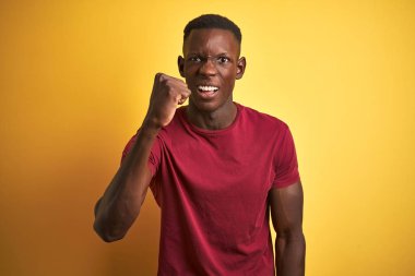 Young african american man wearing red t-shirt standing over isolated yellow background angry and mad raising fist frustrated and furious while shouting with anger. Rage and aggressive concept.