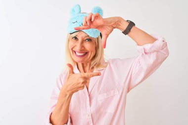 Orta yaş kadın mavi uyku maskesi ve pijama izole beyaz arka plan üzerinde mutlu yüz ile elleri ve parmakları ile çerçeve yapma gülümseyerek giyiyor. Yaratıcılık ve fotoğrafçılık konsepti.