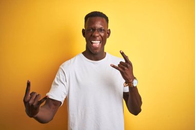 Beyaz tişört giyen genç afro-amerikan adam izole sarı arka plan üzerinde duran deli ifade elleri ile rock sembolü yapıyor bağırıyor. Müzik yıldızı. Ağır konsept.