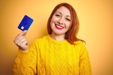 Genç müşteri kızıl saçlı kadın kredi kartını sarı izole edilmiş arka planda tutarken mutlu bir yüzle ayakta duruyor ve gülümsüyor dişlerini gösteren bir gülümsemeyle.