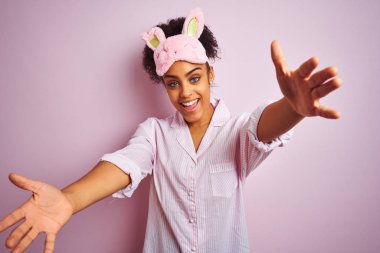 Genç afro-amerikan kadın izole pembe arka plan üzerinde pijama ve maske giyen sarılmak için açık kollarla gülümseyerek kameraya bakıyor. Mutluluğu kucaklayan neşeli ifade.
