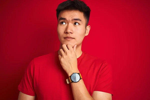 Young asian chinese man wearing  t-shirt standing over isolated red background serious face thinking about question, very confused idea