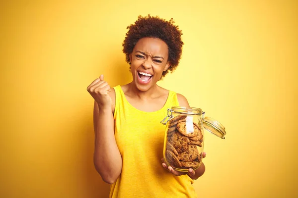 黄色の背景の上にチョコチップクッキーの瓶を持つアフリカ系アメリカ人女性は誇らしげに叫び 勝利と成功を祝う非常に興奮し 感情を応援 — ストック写真