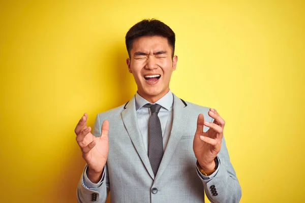 Asian chinese businessman wearing suit and tie standing over isolated yellow background celebrating mad and crazy for success with arms raised and closed eyes screaming excited. Winner concept