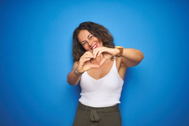 Middle age senior woman with curly hair standing over blue isolated background smiling in love doing heart symbol shape with hands. Romantic concept.