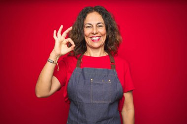 Orta yaş yaşlı kadın kırmızı izole arka plan üzerinde önlük üniforma giyen olumlu el ve parmaklar ile ok işareti yapıyor gülümseyerek. Başarılı ifade.