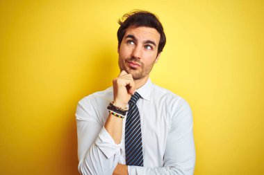 Zarif gömlek giyen ve izole sarı arka plan üzerinde kravat takan genç yakışıklı işadamı, çenesi üzerinde el ile soru, dalgın ifade düşünüyor. Düşünceli bir yüzle gülümsüyor. Şüphe kavramı.