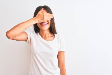 Genç güzel kadın rahat t-shirt izole beyaz arka plan üzerinde ayakta gülümseyerek ve sürpriz için gözleri kaplayan yüzünde el ile gülüyor. Kör kavramı.