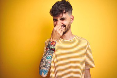 Çizgili tişört giyen genç adam izole sarı arka plan üzerinde duran bir şey kokulu ve, dayanılmaz koku, burun parmakları ile nefes tutarak. Kötü kokular kavramı.